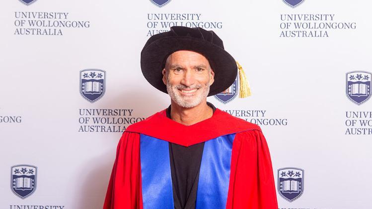 Craig Foster in Honorary Gown in front of white wall with repeated UOW logo
