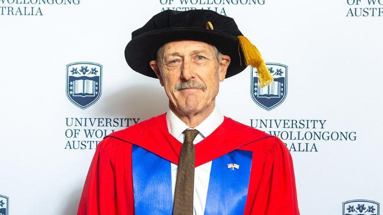 Geoff Morrell in Honorary Gown in front of white wall with repeated UOW logo