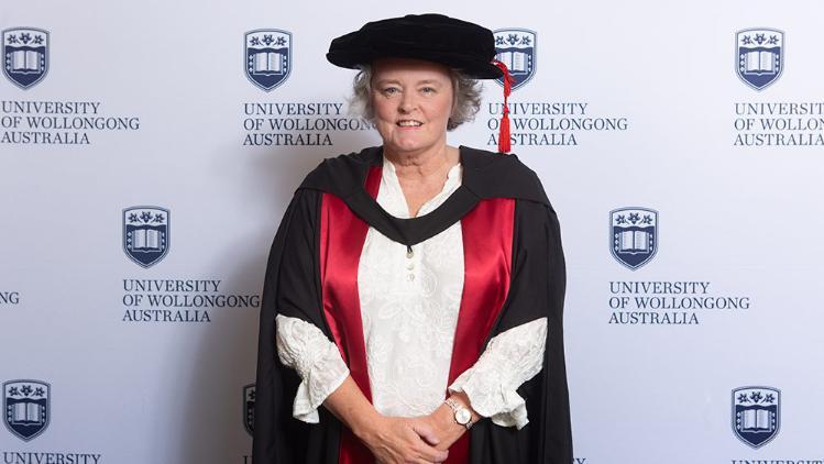 Jennifer Beck in Honorary Gown in front of white wall with repeated UOW logo