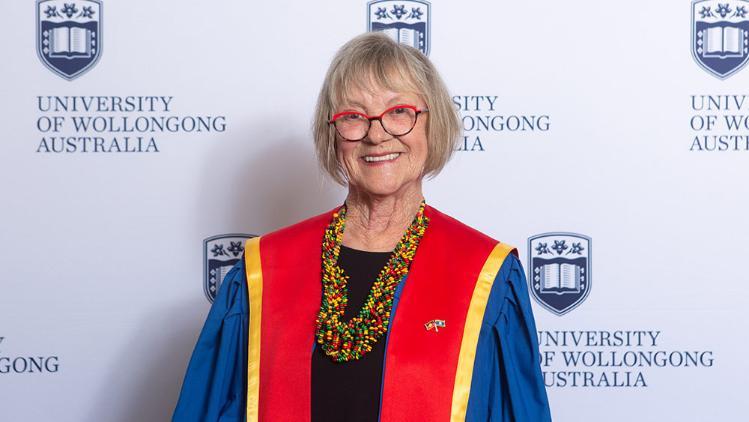 Kay Kent in Honorary Gown in front of white wall with repeated UOW logo