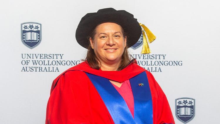 Louise-Sauvage in Honorary Gown in front of white wall with repeated UOW logo